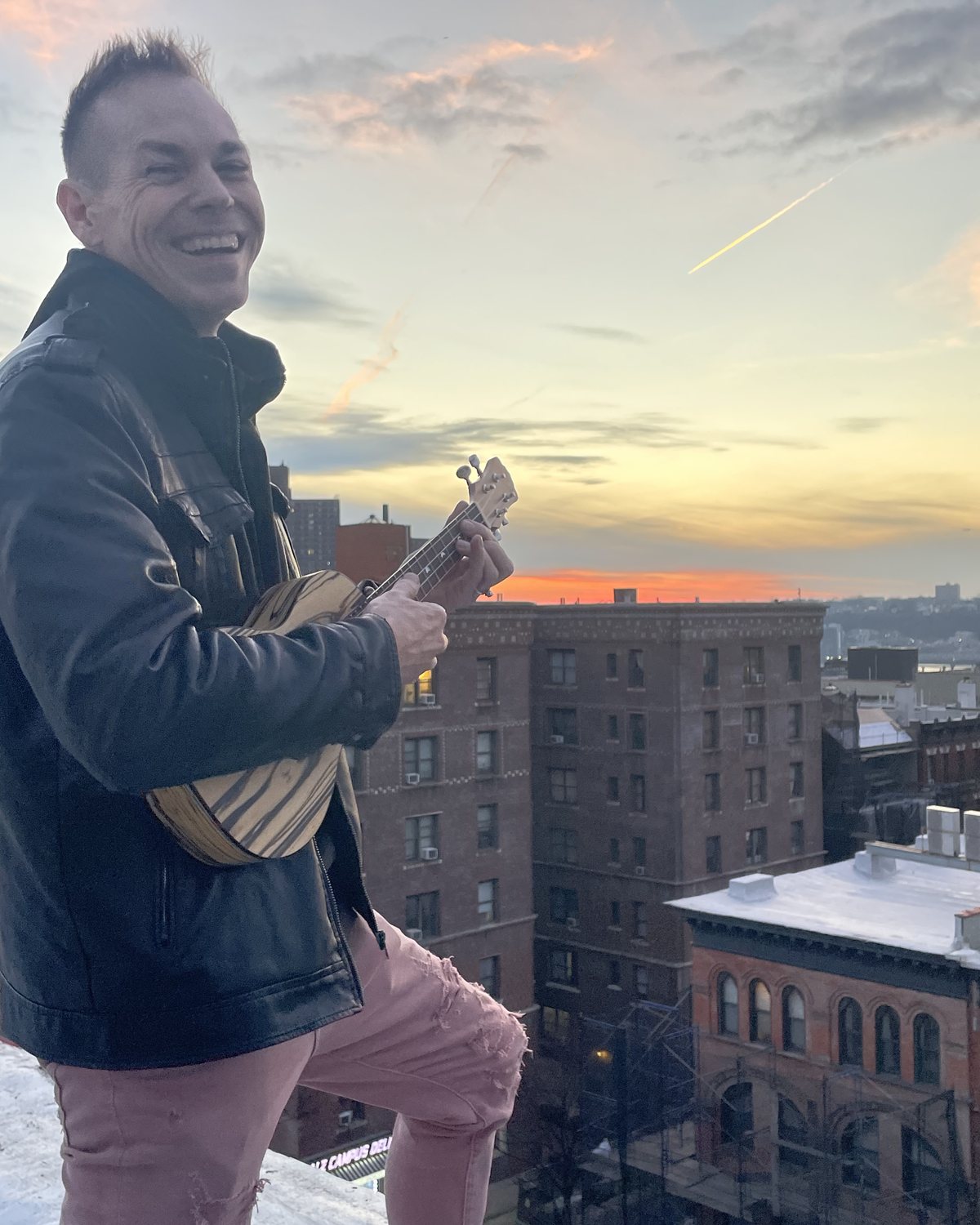Ryan Today laughing and playing a ukulele on a city rooftop at sunset